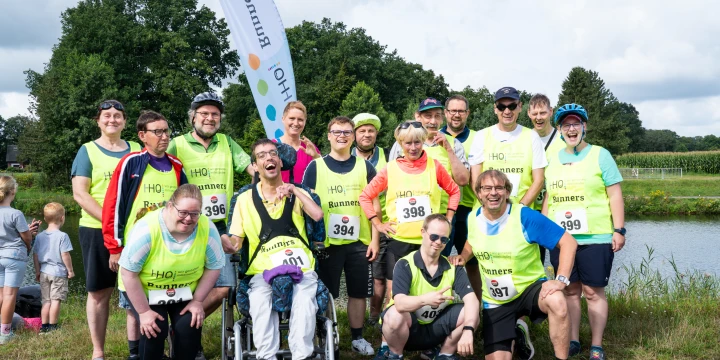 Gruppe von 13 Personen in gelben Sportshirts, teilweise mit Startnummern, posiert vor einer Landschaft mit B&auml;umen, Wasser und Wiese. Im Hintergrund ein wei&szlig;er Banner mit der Aufschrift "HOj Runners". Eine Person sitzt in einem Rollstuhl.