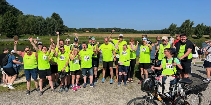Eine Gruppe von etwa 20 Personen in neongelben T-Shirts mit Startnummern, einige jubelnd mit erhobenen Armen. Rechts im Bild eine Person auf einem Fahrrad. Hintergrund: blauem Himmel, B&auml;ume und eine Wiese.