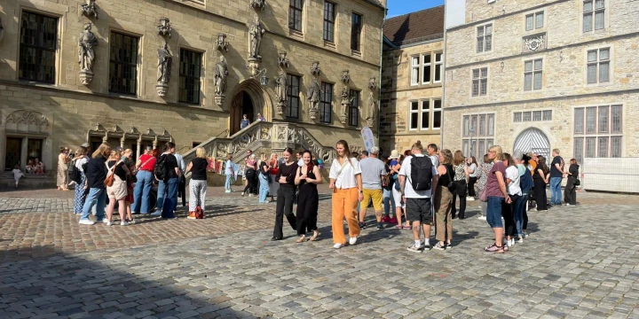 Eine Gruppe von etwa 30 Menschen steht oder geht auf einem gepflasterten Platz vor einem historischen Geb&auml;ude mit Skulpturen an der Fassade und einem steinernen Treppenaufgang. Rechts ein weiteres Geb&auml;ude mit heller Steinfassade und Spitzgiebel. Sonne und Schatten durch klare Wetterbedingungen.