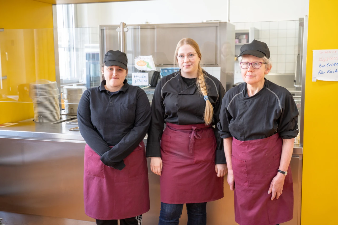 Drei Frauen stehen nebeneinander vor einer Essensausgabe mit gelber Wand. Sie tragen schwarze Kochhemden, schwarze Kopfbedeckungen und bordeauxrote Sch&uuml;rzen. Links gestapelte Teller, im Hintergrund K&uuml;chenutensilien und wei&szlig;e Fliesenwand.