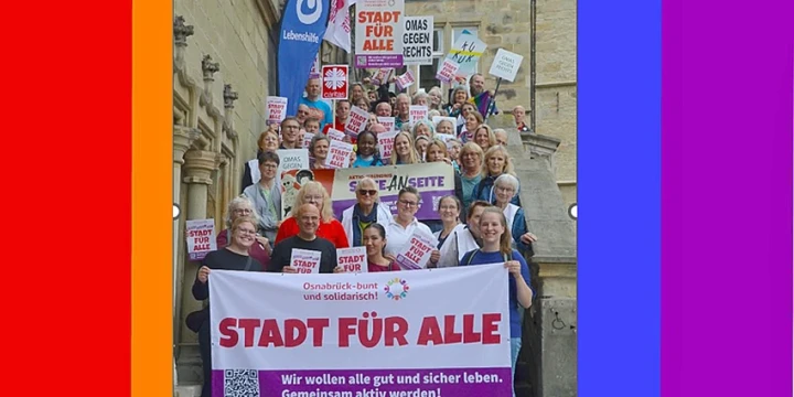 Gruppe von Menschen verschiedenen Alters auf einer Treppe, viele halten Schilder und Banner mit Aussagen wie &bdquo;Stadt f&uuml;r alle&ldquo;, &bdquo;Omas gegen rechts&ldquo; und &bdquo;Osnabr&uuml;ck &ndash; bunt und solidarisch&ldquo;. Hauptbanner mit Text: &bdquo;Stadt f&uuml;r alle. Wir wollen alle gut und sicher leben. Gemeinsam aktiv werden!&ldquo;. Hintergrund zeigt historische Geb&auml;udefassade. Regenbogenfarbiger Rahmen um das Bild.