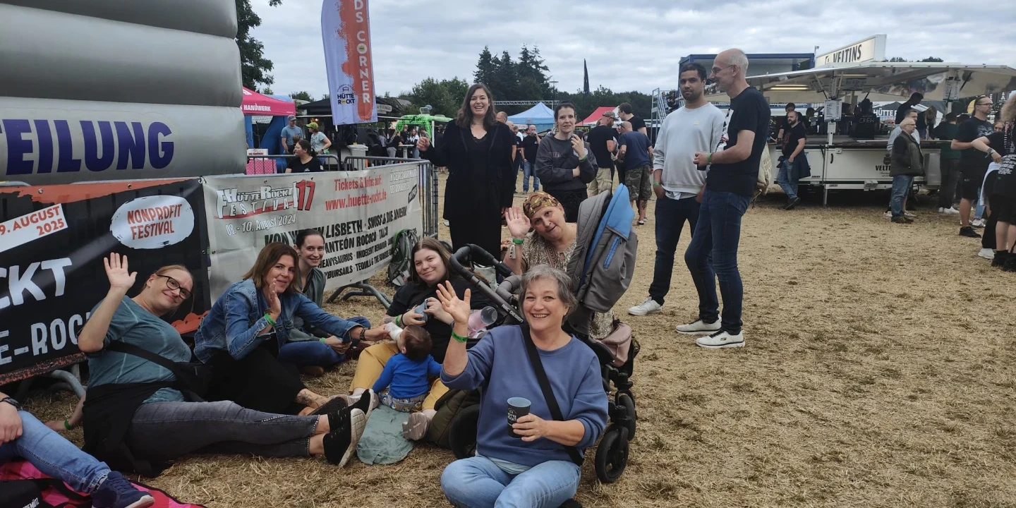 Gruppe von neun Personen unterschiedlichen Alters auf einem Freiluft-Event; sechs davon sitzen oder lehnen entspannt auf trockenem Rasen neben einem Kinderwagen, drei stehen nahe beieinander. Im Hintergrund sind Schilder, St&auml;nde, ein Foodtruck sowie B&auml;ume und bew&ouml;lkter Himmel sichtbar. Ein Banner auf der linken Seite weist auf ein Festival hin.