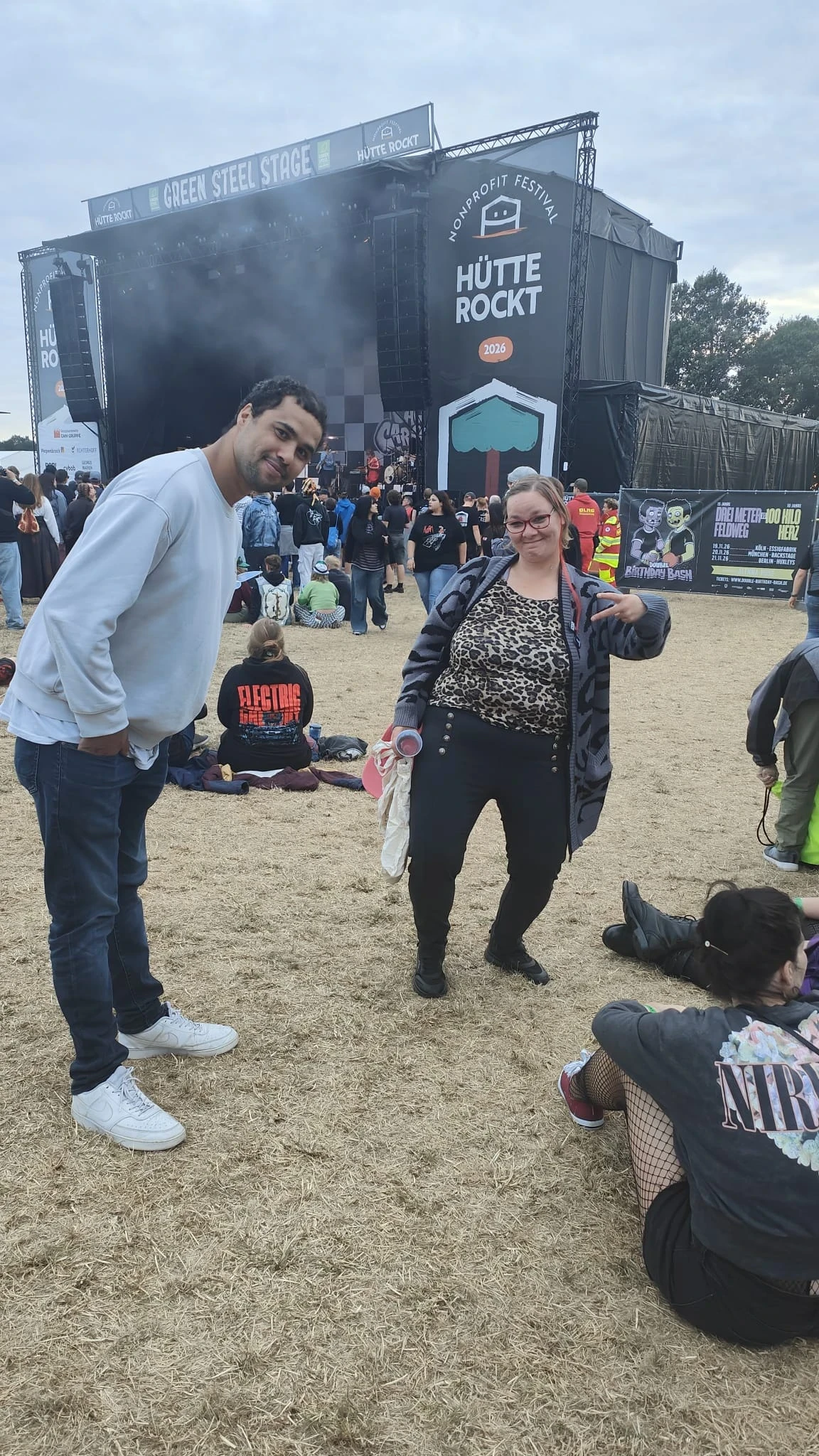 Zwei Personen stehen auf einer trockenen Wiese vor einer gro&szlig;en B&uuml;hne mit der Aufschrift "H&uuml;tte Rockt 2026" und dem Logo des Nonprofit-Festivals "Green Steel Stage". Die Frau tr&auml;gt ein Leopardenmuster-Oberteil, eine graue Jacke und h&auml;lt ein Getr&auml;nk und eine Tasche. Der Mann tr&auml;gt ein wei&szlig;es Sweatshirt und Jeans. Im Hintergrund Menschen, Plakate und sitzende Festivalbesucher.