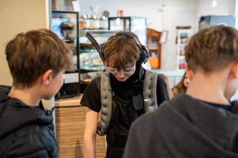 Drei Jugendliche in einem Innenraum. Eine Person in der Mitte tr&auml;gt ein gepolstertes graues Gewichtsweste-&auml;hnliches Kleidungsst&uuml;ck, eine Schutzbrille und Kopfh&ouml;rer. Links und rechts stehen zwei weitere Jugendliche mit dem R&uuml;cken zur Kamera. Im Hintergrund sind ein Tresen mit Glasvitrine und unscharfe Gegenst&auml;nde wie Regale erkennbar.