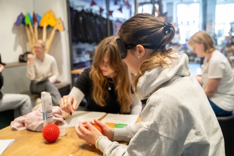 Zwei Frauen sitzen an einem Holztisch in einem Raum mit weiteren Personen im Hintergrund. Eine Frau mit geschnittenem Haar tr&auml;gt eine wei&szlig;e Augenbinde und h&auml;lt einen roten Massageball. Die andere Frau zeigt ihr etwas auf einem Blatt Papier. Auf dem Tisch liegen verschiedene Gegenst&auml;nde, darunter eine Cremeflasche und Stoffe.