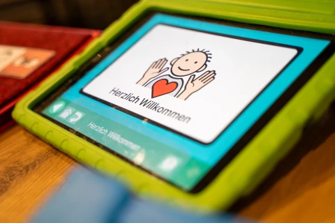 Tablet in gr&uuml;nem Schutzrahmen auf Holztisch, Bildschirm zeigt eine Illustration einer lachenden Figur mit ausgestreckten H&auml;nden, rotem Herz in der Mitte und Text &bdquo;Herzlich Willkommen&ldquo; darunter. Links und rechts neben dem Tablet sind Teile weiterer Objekte sichtbar.