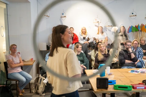 Menschen in einem Raum mit hellen W&auml;nden, sitzend und stehend, einige an Tischen mit Unterrichtsmaterialien. Frauen und M&auml;nner blicken in Richtung einer Frau, die im Vordergrund mit dem R&uuml;cken zur Kamera steht. H&auml;ngelampen und farbige Gegenst&auml;nde im Hintergrund.