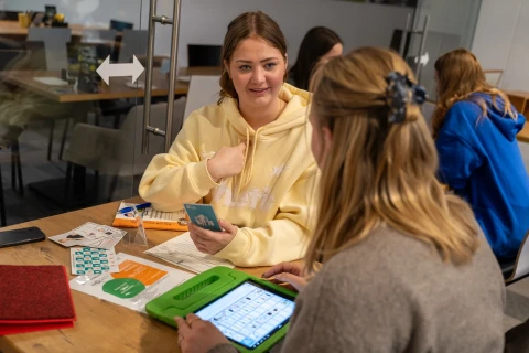 Zwei junge Frauen sitzen an einem Holztisch in einem modernen Arbeitsraum. Die Frau links tr&auml;gt einen gelben Hoodie und h&auml;lt eine kleine Karte. Die Frau rechts tr&auml;gt einen grauen Pullover und hat ein dunkles Haaraccessoire; vor ihr liegt ein gr&uuml;nes Tablet mit einer Tabelle auf dem Bildschirm. Auf dem Tisch liegen weitere Materialien wie Karten, eine rote Mappe und Papierdokumente. Im Hintergrund sind zwei weitere Personen und Glast&uuml;ren zu sehen.