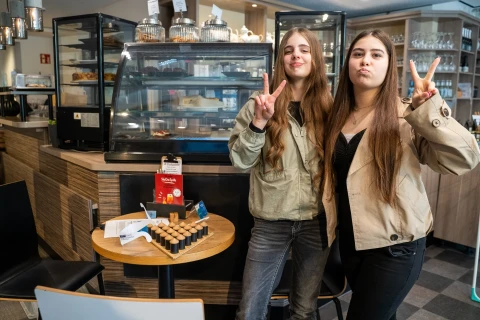 Zwei junge Frauen machen das Victory-Zeichen mit den Fingern, beide tragen lange Haare und helle Jacken. Sie stehen in einem Caf&eacute; vor einer Glastheke mit Geb&auml;ck und anderen Lebensmitteln. Rechts von ihnen ein runder Tisch mit kleinen Flaschen und einem roten Produktkarton. Holz- und Metallelemente dominieren die Einrichtung.