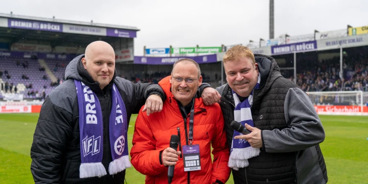 Drei Personen im Fu&szlig;ballstadion, zwei tragen schwarz-graue Kleidung mit lila-wei&szlig;en VfL-Schals, die dritte Person eine rote Jacke. Eine Person h&auml;lt ein Mikrofon. Im Hintergrund sind Spielfeld, Stadiontrib&uuml;nen und Werbebanden erkennbar.