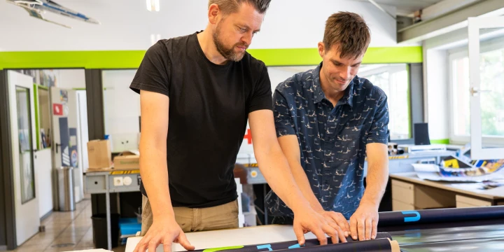 Zwei Personen stehen an einem Tisch in einer Werkstatt mit hellgr&uuml;nen W&auml;nden und gro&szlig;en Fenstern. Sie halten bedruckte Folienrollen in den H&auml;nden, die auf dem Tisch ausgerollt liegen. Im Hintergrund sind Regale, Arbeitsmaterialien und Maschinen erkennbar.