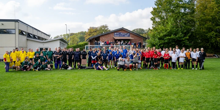 Gruppe von etwa 70 Personen in bunter Sportkleidung, geordnet nach Teams mit gelben, gr&uuml;nen, blauen, roten und wei&szlig;en Trikots, auf einem Fu&szlig;ballfeld. Im Hintergrund ein Ziegelgeb&auml;ude mit Terrasse und Zuschauern, umgeben von B&auml;umen unter leicht bew&ouml;lktem Himmel.