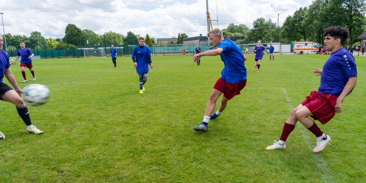 Vier Fu&szlig;ballspieler in roten Shorts und blauen Trikots auf einem gr&uuml;nen Fu&szlig;ballfeld. Ein Spieler schie&szlig;t den Ball, w&auml;hrend andere zuschauen. Im Hintergrund mehrere Spieler, ein Baukran, B&auml;ume und ein geparkter Rettungswagen unter wolkigem Himmel.