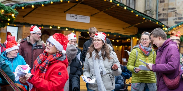 Gruppe von Menschen in Winterkleidung, einige mit roten Weihnachtsm&uuml;tzen, halten Bl&auml;tter in den H&auml;nden, vor einem gelben Holzpavillon mit Lichterkette; Hintergrund: historische Fassade.