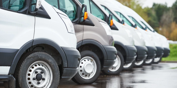 Wei&szlig;e und graue Lieferwagen in einer Reihe geparkt auf nasser Asphaltfl&auml;che, Vorderansichten und Seiten erkennbar, daneben gr&uuml;ne Rasenfl&auml;che und B&auml;ume im Hintergrund.