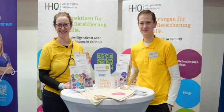 Zwei Personen in gelben Poloshirts stehen an einem hohen wei&szlig;en Stehtisch mit "Karriere Kick"-Logo. Auf dem Tisch liegen Flyer, ein Stoffbeutel und Brosch&uuml;ren. Hinter ihnen drei Rollups der HHO mit Informationsaufschriften wie "Perspektiven f&uuml;r alle" und Themenbereichen wie Pflege, Logop&auml;die, Ergotherapie. Oberhalb ein Schild mit "Leuchtmittel" und "Taschen".