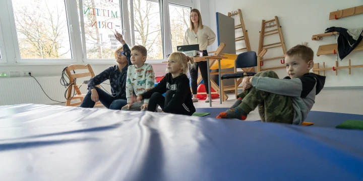 3 Kinder und eine Frau schauen auf eine Leinwand. Im Hintergrund ist ein Projektor zu sehen, an dem eine junge Frau steht.