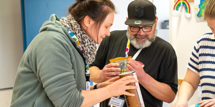 Drei Personen stehen an einem Tisch und basteln gemeinsam. Links eine Person in grauem Oberteil, die eine Pappr&ouml;hre h&auml;lt, in die die mittlere Person gelbe Perlen einf&uuml;llt. Rechts eine Person in gestreiftem Oberteil. Auf dem Tisch bunte Bastelmaterialien wie R&ouml;hren, Perlen, Papier und Dekorationselemente. Im Hintergrund eine blaue T&uuml;r, eine wei&szlig;e Wand und farbige Kinderzeichnungen, darunter ein Regenbogen.