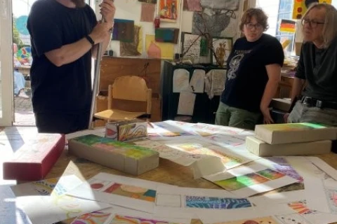Mann mit Glatze und schwarzem T-Shirt h&auml;lt einen zylindrischen Gegenstand in einer Kunstwerkstatt. Auf einem runden Holztisch liegen verschiedene bunte Zeichnungen, Papierb&ouml;gen und flache Pappschachteln. Zwei Frauen beobachten ihn, w&auml;hrend im Hintergrund eine ge&ouml;ffnete Glast&uuml;r und zahlreiche Kunstwerke an der Wand sichtbar sind. Tageslicht f&auml;llt in den Raum.