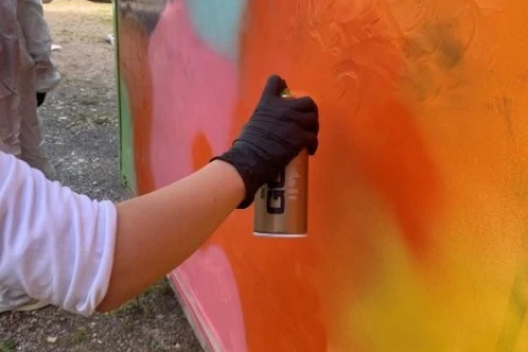 Hand mit schwarzem Handschuh h&auml;lt eine orangefarbene Spr&uuml;hdose und bespr&uuml;ht eine Wand mit leuchtenden Farben, darunter Orange, Rot und Gelb, die ineinander verlaufen. Im Hintergrund ein wei&szlig;er Lastwagen und eine weitere Person mit Spr&uuml;hdose. Boden besteht aus Kieselsteinen.