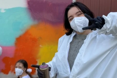 Zwei Personen vor einer wei&szlig;en Wand mit bunten Graffiti-Farbflecken in Blau, Lila, Orange, Rot und Gelb. Beide tragen wei&szlig;e Overalls, schwarzen Handschuhe und FFP2-Masken. Die vordere Person h&auml;lt eine Spr&uuml;hdose und zeigt ein Peace-Zeichen. Links sitzt die zweite Person mit Blick zur Kamera.