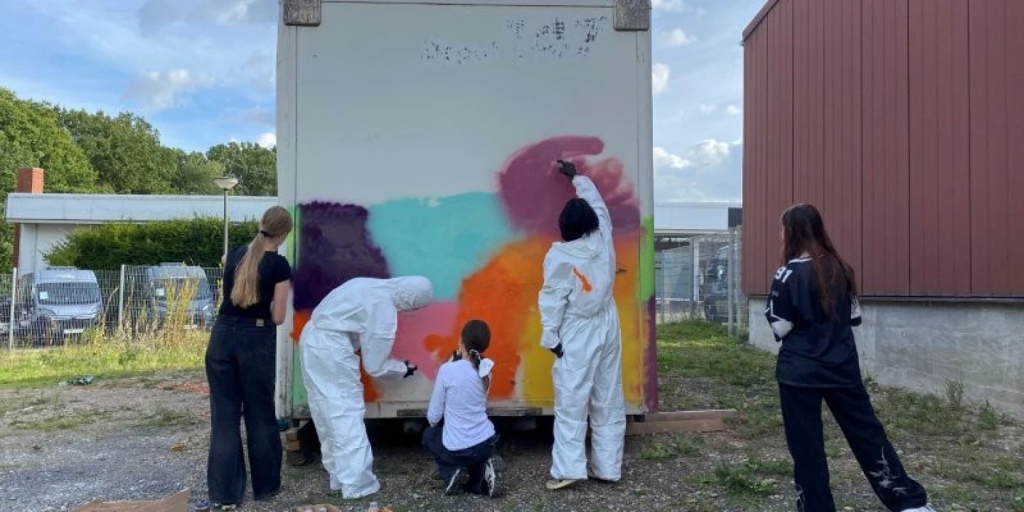 Vier Personen bemalen die R&uuml;ckseite eines wei&szlig;en Containers mit farbigen Spraydosen. Die Farben Rot, Blau, Lila, Orange und Gelb ergeben ein abstraktes Muster. Zwei Personen tragen wei&szlig;e Schutzanz&uuml;ge, eine sitzt, zwei andere stehen. Im Hintergrund sind ein rotes Geb&auml;ude, Z&auml;une, geparkte Autos und B&auml;ume zu sehen, dar&uuml;ber ein bew&ouml;lkter Himmel. Unten links stehen offene Kartons mit Spraydosen.