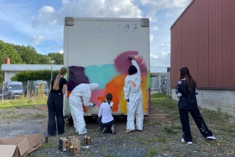 Vier Personen bemalen die R&uuml;ckseite eines wei&szlig;en Containers mit farbigen Spraydosen. Die Farben Rot, Blau, Lila, Orange und Gelb ergeben ein abstraktes Muster. Zwei Personen tragen wei&szlig;e Schutzanz&uuml;ge, eine sitzt, zwei andere stehen. Im Hintergrund sind ein rotes Geb&auml;ude, Z&auml;une, geparkte Autos und B&auml;ume zu sehen, dar&uuml;ber ein bew&ouml;lkter Himmel. Unten links stehen offene Kartons mit Spraydosen.