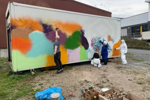 Personen bemalen einen wei&szlig;en Container mit Spraydosen. Linke Wandh&auml;lfte bereits mit bunten Farbfl&auml;chen in Orange, Gr&uuml;n, Lila und Blau bedeckt. Vier Personen, zwei in wei&szlig;en Schutzanz&uuml;gen. Boden mit Spraydosen, Planen und Zubeh&ouml;r bedeckt. Geb&auml;udeteile im Hintergrund.