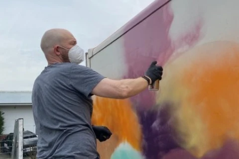 Mann mit grauem T-Shirt, schwarzer Hose, schwarzen Handschuhen und wei&szlig;er Atemschutzmaske spr&uuml;ht mit einer Spraydose bunte Farben (Orange, Lila, Gelb, T&uuml;rkis) auf eine helle Wand. Rechts hinter ihm eine Stufenleiter, links Zaun und Gr&uuml;nfl&auml;che. Himmel bew&ouml;lkt.