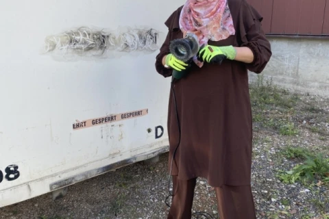 Person in brauner Kleidung und buntem Kopftuch, tr&auml;gt Schutzbrille, Mundschutz und gr&uuml;ne Arbeitshandschuhe, h&auml;lt ein Werkzeug mit Kabel vor einem wei&szlig;en Container mit Schriftzug &bdquo;Erst gesperrt gesperrt&ldquo;. Untergrund aus Kies, links Grasb&uuml;schel, rechts Wandabschnitt.