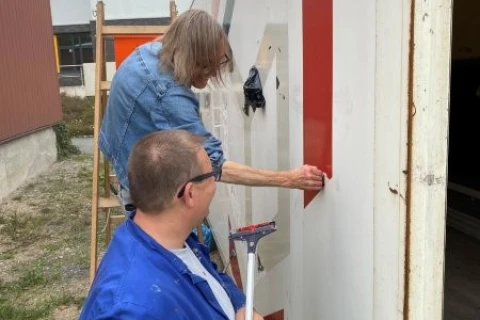 Zwei Personen arbeiten an der Seite eines wei&szlig;en Fahrzeugs mit gro&szlig;en roten Buchstaben. Die vordere Person tr&auml;gt eine blaue Arbeitsjacke, h&auml;lt ein Werkzeug und schaut nach oben. Die zweite Person steht auf einer Leiter und bringt eine rote Folie auf die Fahrzeugseite an. Im Hintergrund sind ein rotes Geb&auml;ude, ein weiteres Geb&auml;ude mit grauem Dach und ein grasbewachsener Boden sichtbar.