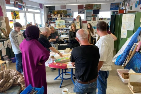 Gruppe von acht Personen in einem hell beleuchteten Kunst- oder Kreativraum, versammelt um einen runden, mit Materialien wie Papier und Werkzeugen bedeckten Tisch. An den W&auml;nden zahlreiche Zeichnungen, Gem&auml;lde und Regale mit Farben und Utensilien.
