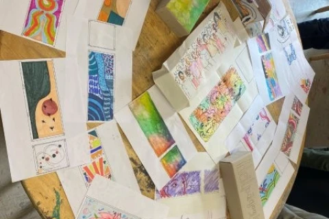 Verschiedene farbenfrohe Zeichnungen und Karten, gr&ouml;&szlig;tenteils rechteckig, sind auf einem runden Holztisch ausgebreitet. Die Motive umfassen abstrakte Muster, Blumen, geometrische Formen und Regenbogenfarben. Einige Karten sind aufgerichtet, andere liegen flach. Am Rand des Tisches sind mehrere Personen, die teilweise sichtbar sind.