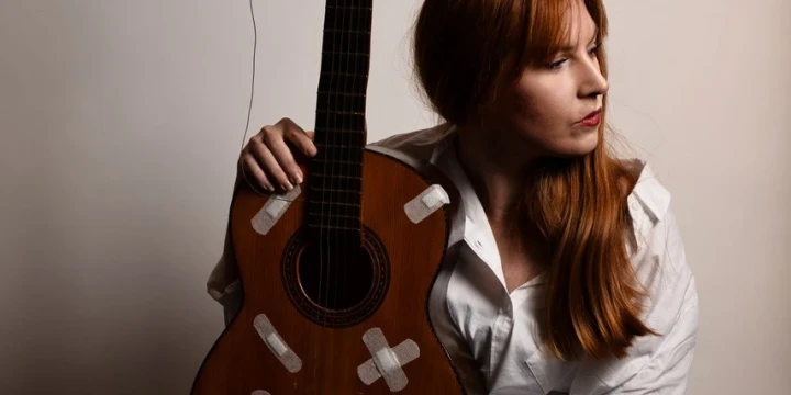 Frau mit langen roten Haaren und wei&szlig;er Bluse sitzt und lehnt eine klassische braune Akustikgitarre mit zahlreichen wei&szlig;en Pflastern darauf an ihre Schulter. Ein d&uuml;nnes Kabel ist an die Gitarre angeschlossen. Der Hintergrund ist beige und gleichm&auml;&szlig;ig.