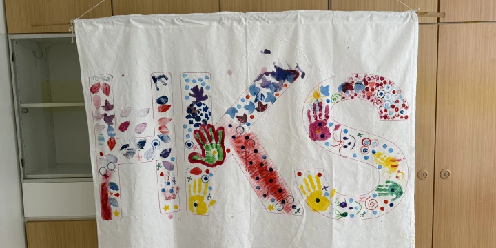 Wei&szlig;e Stoffbahn mit bemalten Gro&szlig;buchstaben "HKS", dekoriert mit farbigen Handabdr&uuml;cken, Punkten, Strichen und Mustern in verschiedenen Farben wie Rot, Blau, Gelb und Gr&uuml;n. Der Stoff ist an einer Leine vor Holzschr&auml;nken aufgeh&auml;ngt.