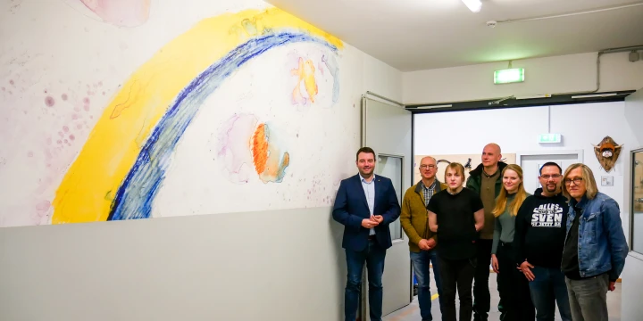 Eine Gruppe von sechs Personen steht in einem Flur vor einer gro&szlig;en, abstrakten Wandmalerei. Die Malerei besteht aus einem geschwungenen gelben und blauen Streifen sowie farbigen, diffusen Formen auf wei&szlig;em Hintergrund. Im Flur sind wei&szlig;e W&auml;nde, eine T&uuml;r mit gr&uuml;nem Notausgangsschild und ein brauner Boden erkennbar.
