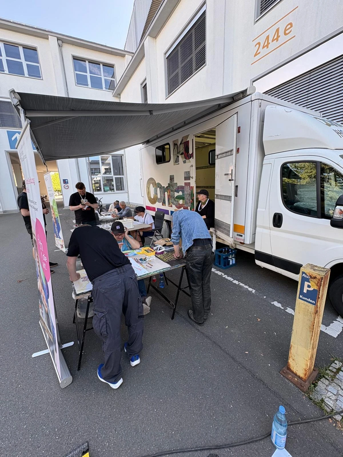 Wei&szlig;er Transporter mit offener Seitenklappe und aufgespannter Markise, darunter mehrere Tische mit Personen, die an Bastelmaterialien arbeiten. Schilder und Plakate stehen links, Geb&auml;ude mit gro&szlig;en Fenstern im Hintergrund.