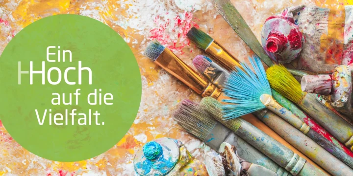 Gr&uuml;ne Kreisfl&auml;che links mit wei&szlig;em Text: "Ein Hoch auf die Vielfalt." Rechts daneben eine Ansammlung bunter, mit Farbe verschmierter Pinsel unterschiedlicher Gr&ouml;&szlig;e und Form, liegend auf einer unregelm&auml;&szlig;ig bemalten, gelb-orangenen Unterlage. Farbtuben mit Farbresten sind zwischen den Pinseln erkennbar.