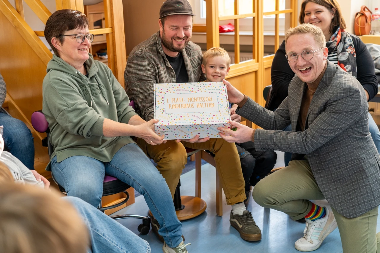 Eine Person in gr&uuml;ner Kapuzenjacke und eine Person in grauem Blazer halten gemeinsam eine rechteckige, wei&szlig;e Schachtel mit bunter Aufschrift "1. Platz: Montessori Kindergarten Wut&ouml;schingen". Sie sitzen in einem Raum mit Holzm&ouml;beln und blauen Bodenfliesen, umgeben von weiteren Personen.