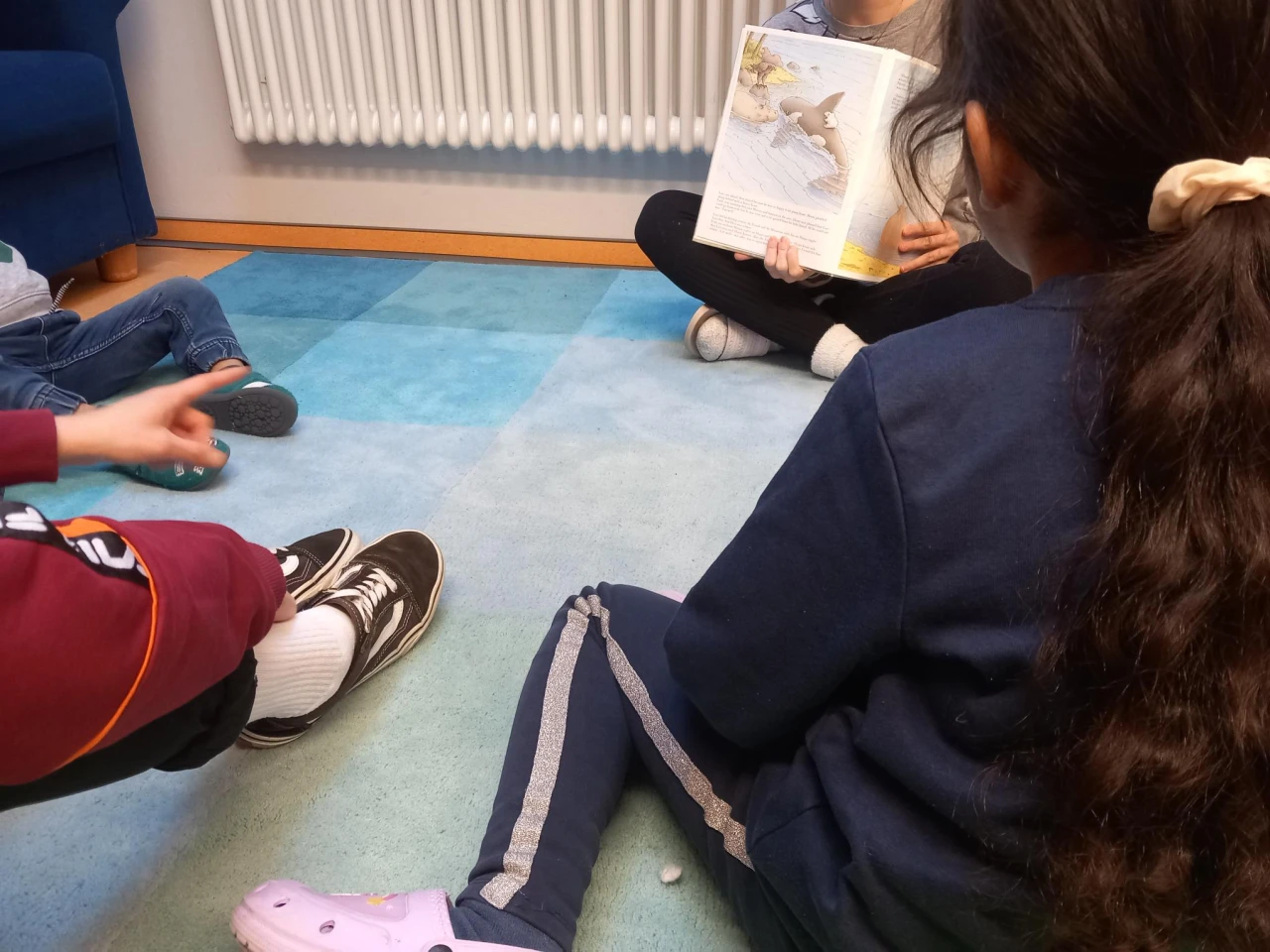 Kinder sitzen im Kreis auf einem blau gemusterten Teppich. Eine Person mit einem aufgeschlagenen Bilderbuch zeigt eine Illustration. Linke Bildseite: ausgestreckte Hand eines Kindes. Rechte Seite: Kind mit dunklen Haaren und dunkelblauer Kleidung. Hintergrund: wei&szlig;er Heizk&ouml;rper und blaue Couch.
