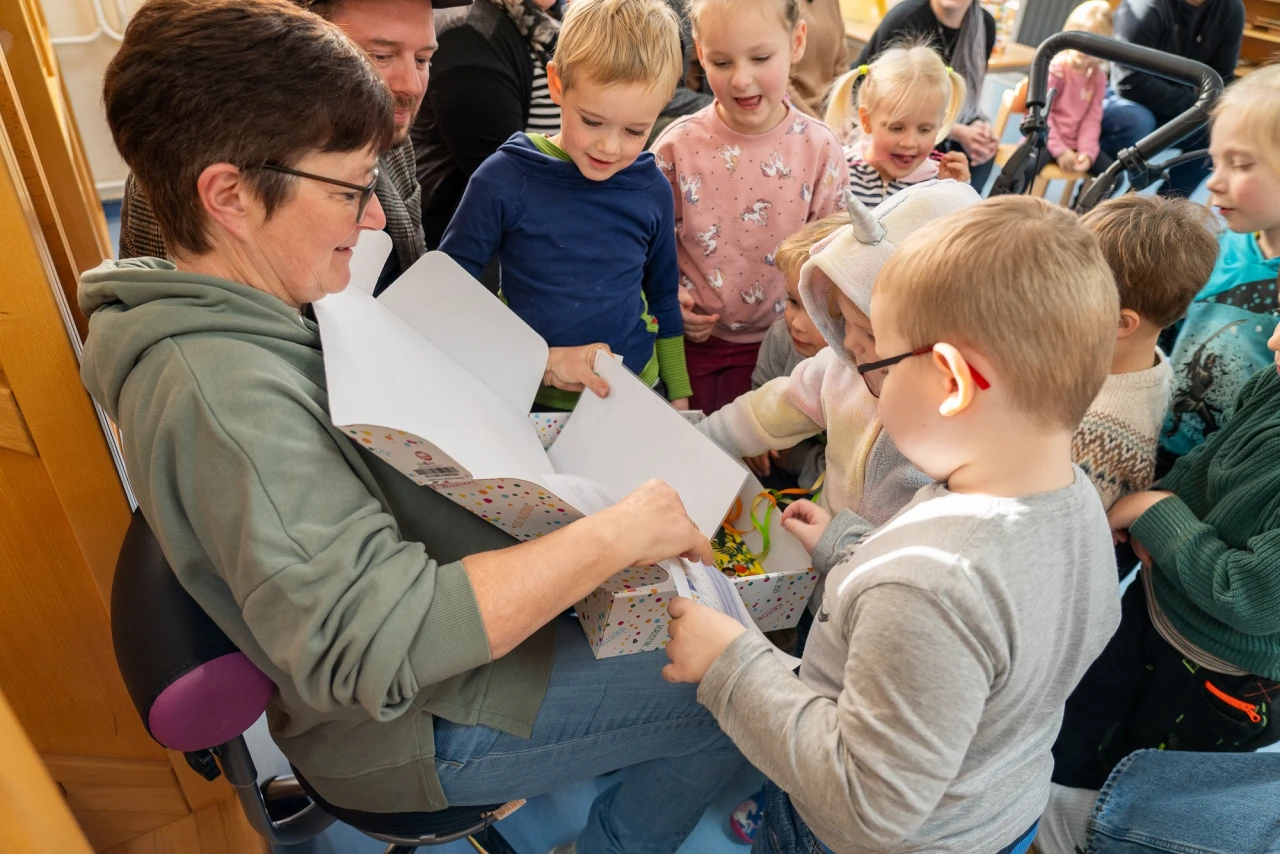 Eine Gruppe von Kindern umgibt sitzende erwachsene Person in gr&uuml;nem Kapuzenpullover, die eine ge&ouml;ffnete rechteckige Geschenkbox mit buntem Muster h&auml;lt. In der Box befinden sich S&uuml;&szlig;igkeiten oder kleine Gegenst&auml;nde. Die Kinder tragen unterschiedliche Kleidung und blicken interessiert auf die Box.