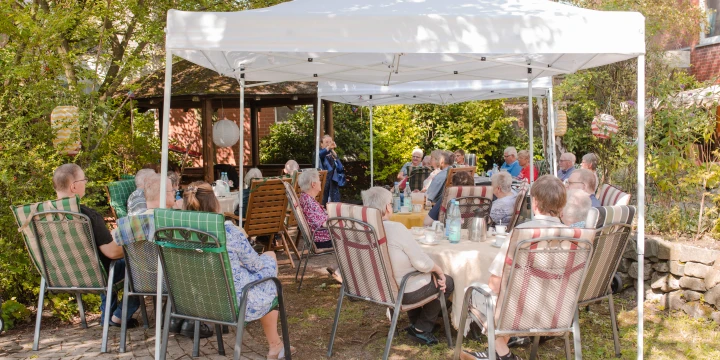 Menschen unterschiedlichen Alters sitzen auf St&uuml;hlen an mehreren Tischen unter einem wei&szlig;en Pavillon im Freien. Die Tische sind mit Tischdecken, Wasserflaschen und Gl&auml;sern gedeckt. Im Hintergrund B&auml;ume, Str&auml;ucher, ein Backsteingeb&auml;ude und eine steinerne Gartenmauer. Links hinter dem Pavillon steht eine Person und spricht.