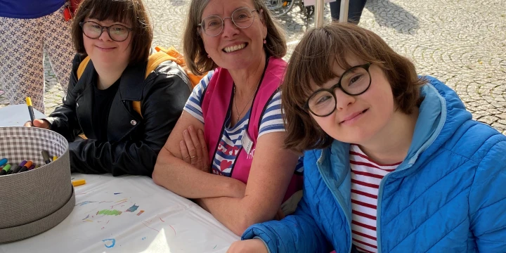 Ein Tisch mit einer wei&szlig;en Tischdecke, darauf Holzbausteine, Stifte, eine Wasserflasche und eine Stiftdose. Drei Personen in bunter Kleidung sind am Tisch sitzend. Im Hintergrund ein Marktplatz mit Kopfsteinpflaster, mehreren stehenden Personen, einem Rollstuhl, Schildern und historischen Geb&auml;uden mit gro&szlig;en Fenstern.