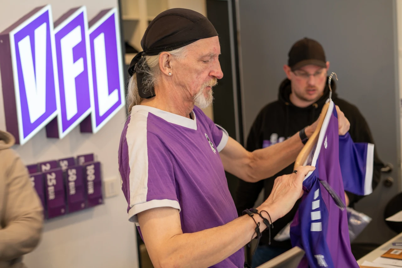 Person mit grauem Haar in einem lila Trikot mit wei&szlig;en Akzenten h&auml;lt ein weiteres lila Trikot auf einem B&uuml;gel. Im Hintergrund lila VFL-Logo und Preisschilder f&uuml;r Merchandise-Artikel.