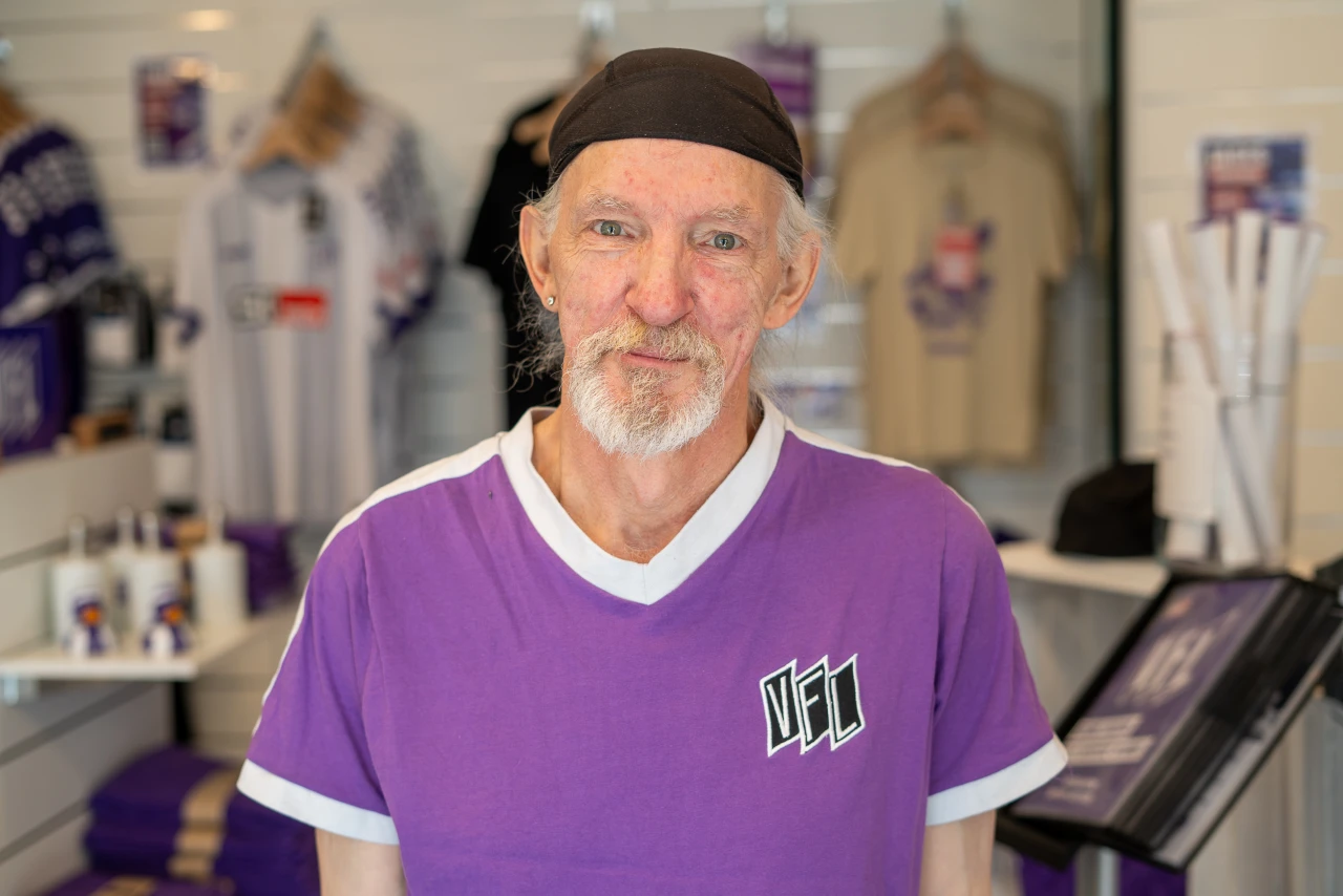 Person in lila T-Shirt mit wei&szlig;em Kragen und VfL-Logo, im Hintergrund Verkaufsregale mit aufgeh&auml;ngten Fu&szlig;balltrikots, Merchandise-Artikeln und weiteren Waren.