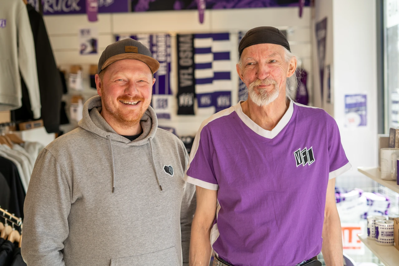 Zwei Personen stehen in einem Fanartikel-Shop mit lila-wei&szlig;er Dekoration. Die linke Person tr&auml;gt einen grauen Kapuzenpullover und eine graue Kappe. Die rechte Person tr&auml;gt ein lila T-Shirt mit wei&szlig;em Kragen und dem Logo des VfL. Im Hintergrund sind Regale mit Fanartikeln, darunter Schals und T-Shirts, zu sehen.