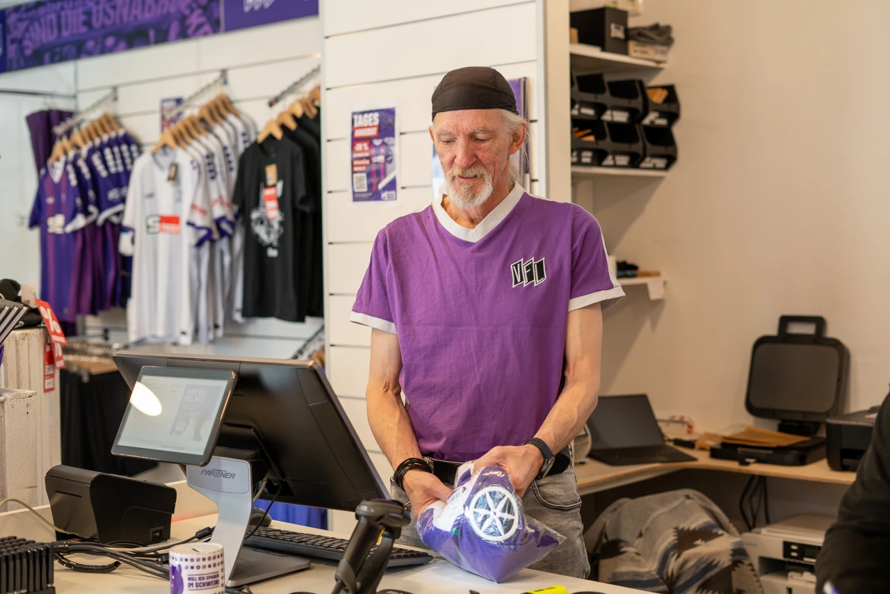 Ein Verk&auml;ufer in einem Fanshop tr&auml;gt ein lila T-Shirt mit der Aufschrift &bdquo;VE1&ldquo; und h&auml;lt einen lila Trikotbeutel mit einem wei&szlig;en Logo. Im Hintergrund helle Verkaufsregale mit Sportkleidung, darunter lila und wei&szlig;e Trikots, sowie weitere Fanartikel. Links eine Registrierkasse auf einem Tresen, rechts technisches Equipment.
