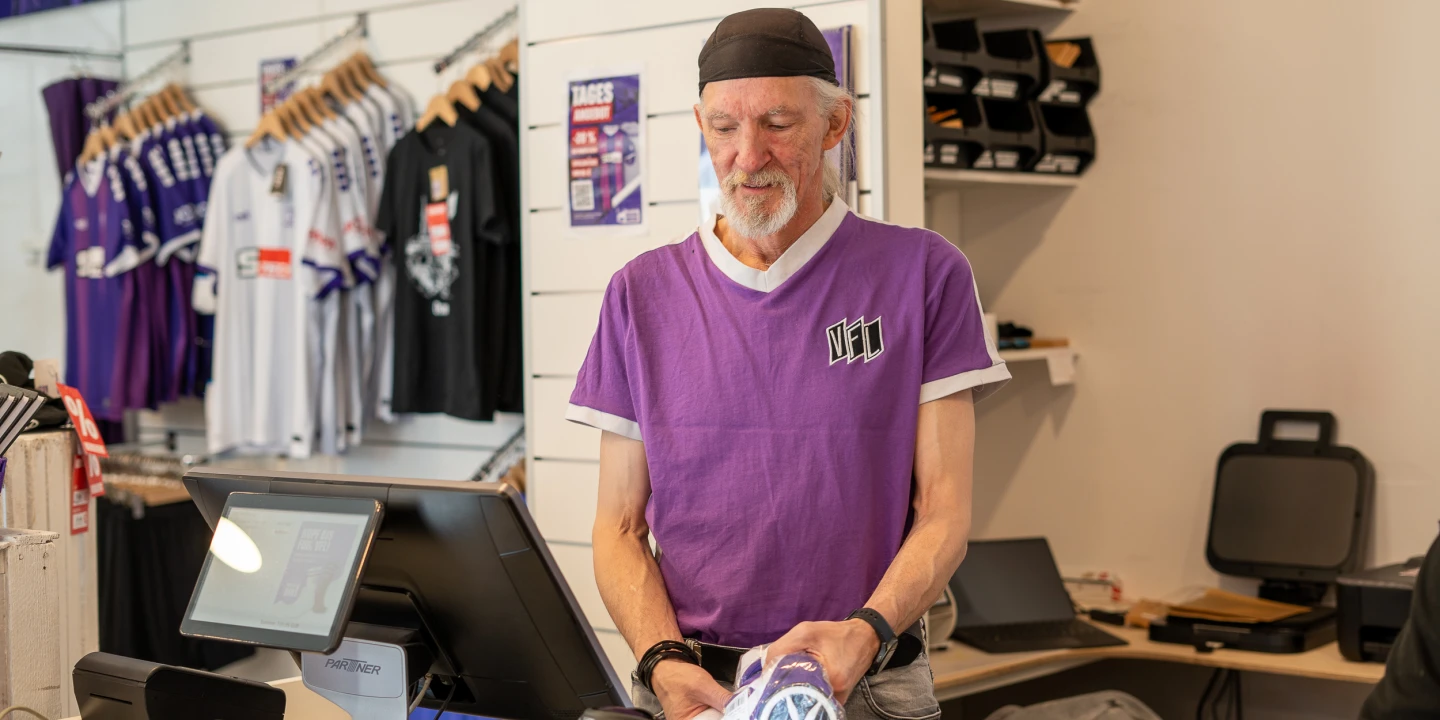 Ein Verk&auml;ufer in einem Fanshop tr&auml;gt ein lila T-Shirt mit der Aufschrift &bdquo;VE1&ldquo; und h&auml;lt einen lila Trikotbeutel mit einem wei&szlig;en Logo. Im Hintergrund helle Verkaufsregale mit Sportkleidung, darunter lila und wei&szlig;e Trikots, sowie weitere Fanartikel. Links eine Registrierkasse auf einem Tresen, rechts technisches Equipment.