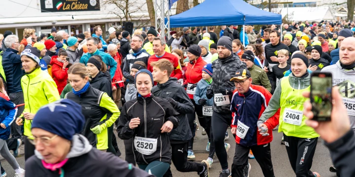 Menschen verschiedener Altersgruppen in Sportbekleidung laufen w&auml;hrend eines Wettlaufs eine Stra&szlig;e entlang. &Uuml;ber ihnen ein blaues Banner mit der Aufschrift "Ziel". Im Hintergrund Zuschauer, ein Geb&auml;ude mit Restaurant "La Chiesa" und ein blauer Pavillon.