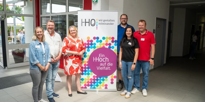 Sechs Personen stehen nebeneinander in einem Innenraum vor einem Roll-up-Poster. Das Poster zeigt bunte Kreispunkte und den Text: "HHO - wir gestalten miteinander. Ein HOOch auf die Vielfalt." Links Glasfront und Au&szlig;enbereich, rechts Flur mit T&uuml;r. Grauer Fliesenboden.